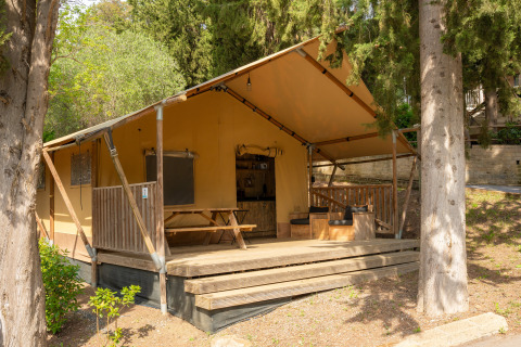 Safari-Zelt mit Holzterrasse im Grünen, Sitzbereich im Freien und Picknicktisch unter Bäumen.