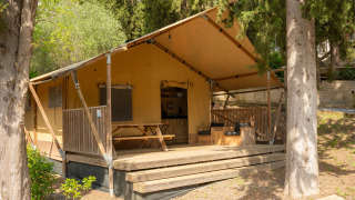 Safari-Zelt mit Holzterrasse im Grünen, Sitzbereich im Freien und Picknicktisch unter Bäumen.