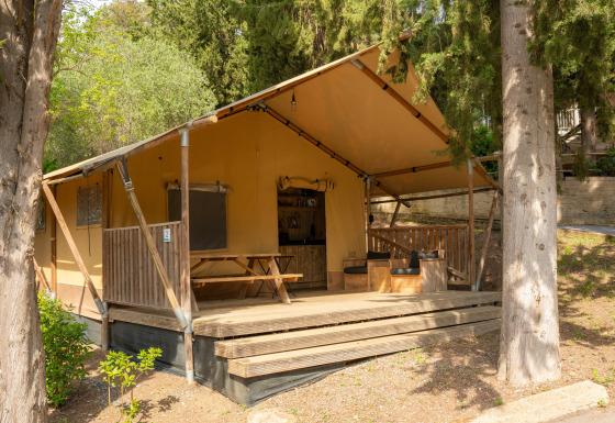 Tente safari avec terrasse en bois, coin salon extérieur et table de pique-nique entourée d’arbres.
