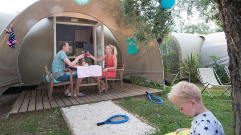 Gezin geniet van een maaltijd bij een Coco Suite safaritent, kind speelt op het gras in de zomer.