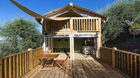 Safari tent Air lodge met houten terras, buitentafel en stoelen, gelegen tussen bomen en blauwe lucht.