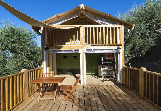 Safari tent Air lodge met houten terras, buitentafel en stoelen, gelegen tussen bomen en blauwe lucht.