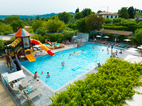 Holiday park pool area with water slides, people swimming and relaxing, surrounded by greenery in Lombardy, Italy.
