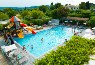 Holiday park pool area with water slides, people swimming and relaxing, surrounded by greenery in Lombardy, Italy.