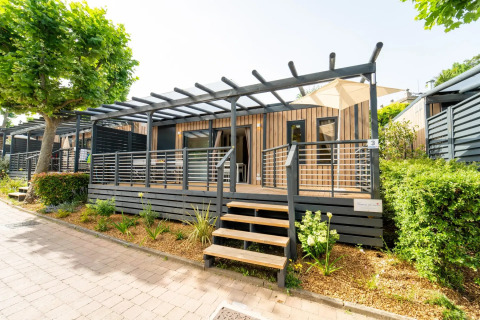 Lodge moderno con terraza amplia, barandilla de madera y vegetación alrededor, bajo el sol brillante.