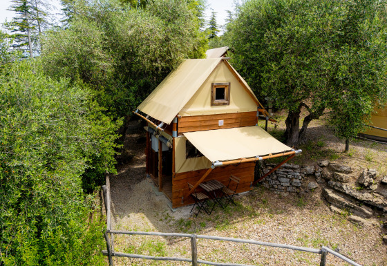 La tente safari Air Stay XL se trouve parmi les arbres, avec terrasse et petit jardin clôturé en bois.