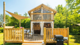 Airlodge 2.0, casetta di legno con ampia veranda, barbecue e sedie, immersa nella natura verde e luminosa.