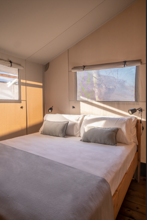 Slaapkamer in een safaritent met comfortabel bed, kussens en zonlicht dat via de ramen naar binnen valt.