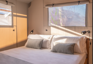 Slaapkamer in een safaritent met comfortabel bed, kussens en zonlicht dat via de ramen naar binnen valt.
