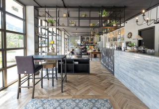 Modern café interior with tall windows, stylish furniture, and lots of plants at Lago Idro Glamping Boutique, Italy.