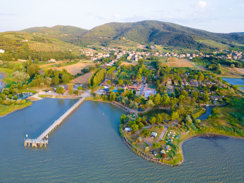 Vista aerea di Trasimeno Glamping Resort in Umbria, Italia, con lago, pontile, alberi e colline sullo sfondo.