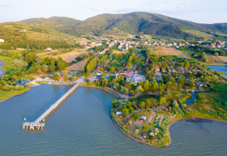 Aerial view of Trasimeno Glamping Resort in Umbria, Italy, showing lake, pier, trees, and surrounding hills.