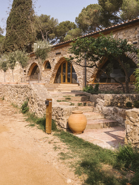 Edificio in pietra con archi e porta in legno tra alberi e piante a Wecamp Cala Montgó, Catalogna, Spagna.