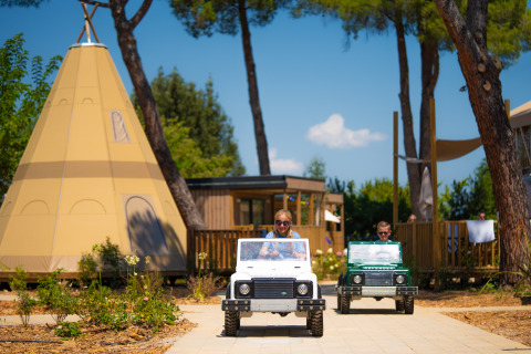 Children driving mini electric cars in front of teepees and lodges at Trasimeno Glamping Resort in Umbria, Italy.