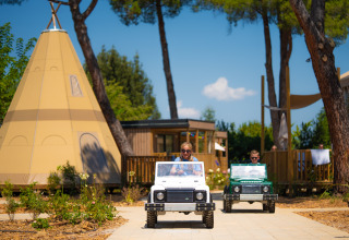 Children driving mini electric cars in front of teepees and lodges at Trasimeno Glamping Resort in Umbria, Italy.