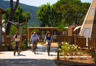 Tre persone vanno in bicicletta su un sentiero a Trasimeno Glamping Resort, circondati da lodge e vegetazione.