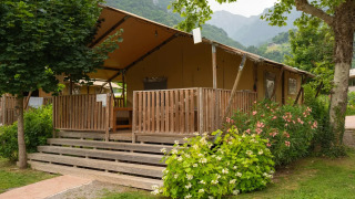 Safari-Zelt mit Holzveranda, von grünen Bäumen und Blumen umgeben, in einer bergigen Landschaft.