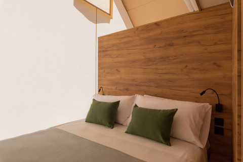 Gezellig bed in een safaritent met houten hoofdbord, witte lakens en groene kussens in zacht licht.