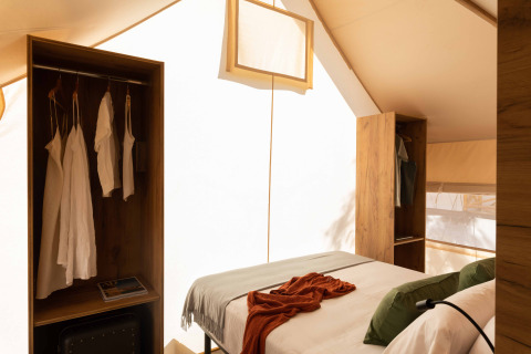 Interieur van een safaritent met comfortabel bed, open kasten en zacht natuurlijk licht.