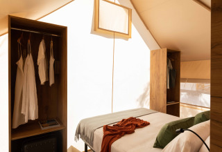 Interieur van een safaritent met comfortabel bed, open kasten en zacht natuurlijk licht.