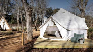 Safari-Zelt namens Bell Tent auf Holzterrasse im Wald, eingerichtet mit Bett und moderner Außensitzgelegenheit.