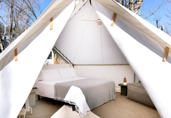 Vista interna di una tenda safari con letto matrimoniale, comodini e alberi ben visibili all'esterno.