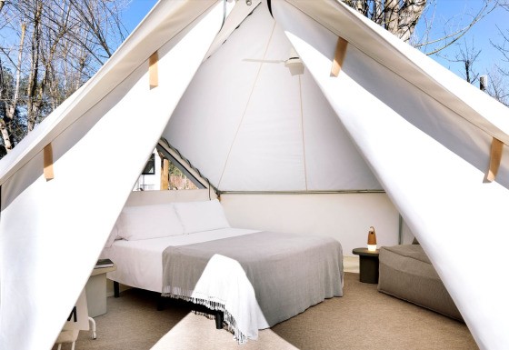 Vista interna di una tenda safari con letto matrimoniale, comodini e alberi ben visibili all'esterno.