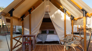 Tienda safari Glamping Couple con piso de madera, cama doble, mesa exterior, sillas y lámpara en la entrada.