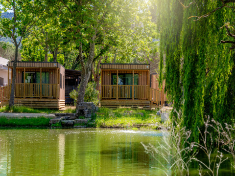 Dos cabañas de madera frente al lago en Cottage Next XL Loggia Laguna, rodeadas de árboles y luz solar.