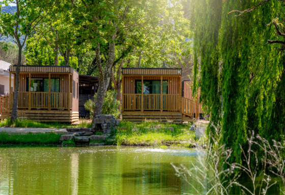 Dos cabañas de madera frente al lago en Cottage Next XL Loggia Laguna, rodeadas de árboles y luz solar.