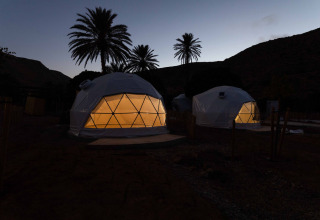 Verlichte geodetische koepeltenten bij schemering in Wecamp Cabo de Gata, Spanje, tussen palmbomen.