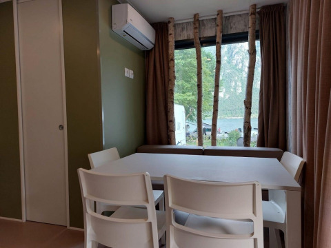 Comedor interior con sillas blancas, aire acondicionado y gran ventana con vista al lago y árboles.