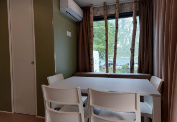 Comedor interior con sillas blancas, aire acondicionado y gran ventana con vista al lago y árboles.