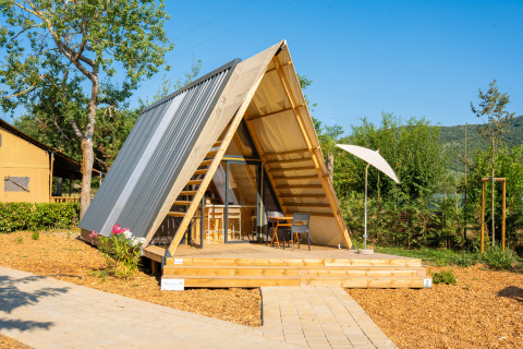Luxe A-frame safaritent met houten terras en stoelen, omgeven door groene natuur bij zonnig weer.