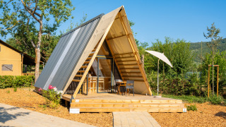 Tenda safari di lusso a forma di A con terrazza in legno e sedie, circondata dalla natura al sole.