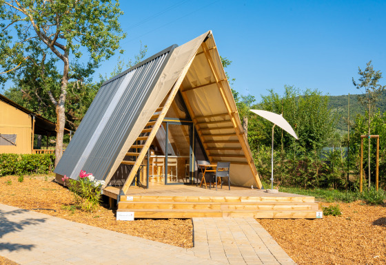 Luxe A-frame safaritent met houten terras en stoelen, omgeven door groene natuur bij zonnig weer.