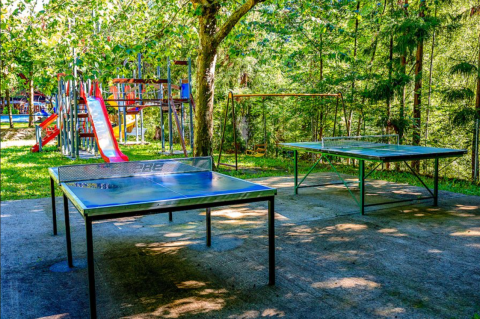 Outdoor playground at a campsite with two table tennis tables, slides, and swings surrounded by greenery.
