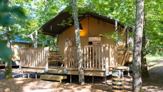 Tenda safari nei boschi con terrazza rialzata in legno, staccionata e tavolo, circondata da alberi verdi.
