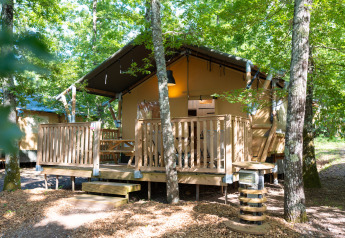Safari-Zelt im Wald mit Holzterrasse, Holzzaun und Picknickbank, umgeben von grünen Bäumen.