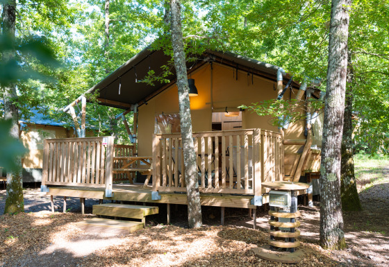 Tente safari dans les bois avec terrasse en bois surélevée, rambarde et table, entourée d’arbres verts.