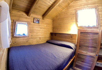 Cozy wooden cabin with a double bed, blue quilt, and two small windows at Campsite Bedura Park, Spain.
