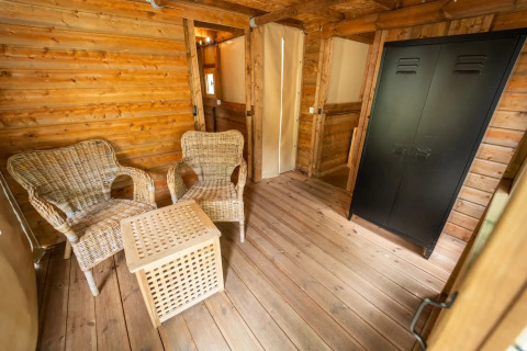 Intérieur d'une tente safari avec deux fauteuils en osier, une table et une armoire noire sur plancher en bois.