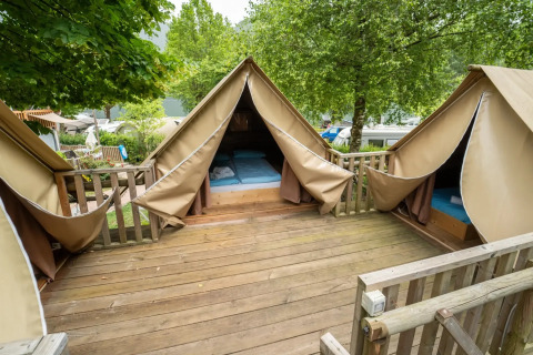 Tienda safari con terraza de madera y tres áreas para dormir, rodeada de árboles y naturaleza verde.