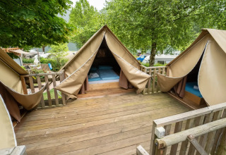 Tienda safari con terraza de madera y tres áreas para dormir, rodeada de árboles y naturaleza verde.