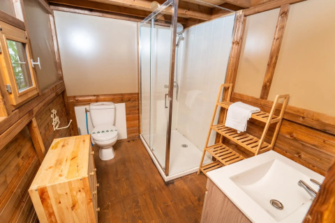 Salle de bain dans une tente safari avec panneaux de bois, douche, toilettes, lavabo et étagère à serviettes.