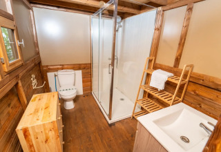 Salle de bain dans une tente safari avec panneaux de bois, douche, toilettes, lavabo et étagère à serviettes.