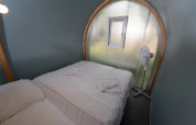 Glampingkamer Coco Couple met een tweepersoonsbed, witte lakens, klein raam en een ventilator.