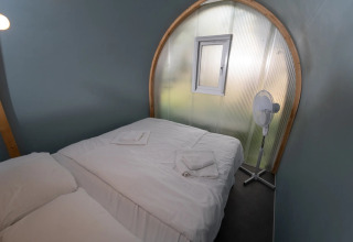 Et glampingværelse i Coco Couple med dobbeltseng, hvidt sengetøj, lille vindue og en ventilator.