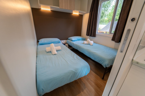 Chambre glamping avec deux lits simples, draps bleus, serviettes blanches et fenêtre donnant sur l’extérieur.