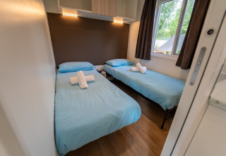 Chambre glamping avec deux lits simples, draps bleus, serviettes blanches et fenêtre donnant sur l’extérieur.